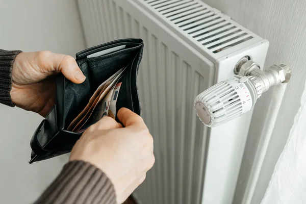 Adamın biri içinde para dolu banknotlar olan bir cüzdanı ısıtma kaloriferinin yanında tutuyor. Isıtma ve doğalgaz fiyatları yükseliyor. Avrupa 'da Enerji Krizi.