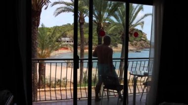 Siluetli beyaz bir adamın bir otel balkonuna çıkıp güneşli bir yaz gününde İspanya 'nın İbiza adasında rahatlamak için oturduğu görüntüler.