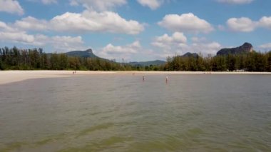 The beautiful beach in the town of Krabi Ao Nang Beach in Thailand, showing people walking on the beach and having fun in the ocean with white fluffy clouds in the sky.