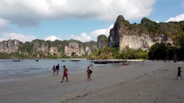 People relaxing on the beach of Railay Beach in AO Nang in Thailand, taken on a hot sunny say with a few clouds in the sky with people on the beach and long tail boats.