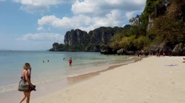 People relaxing on the beach of Railay Beach in AO Nang in Thailand, taken on a hot sunny say with a few clouds in the sky with people on the beach near the mountains.