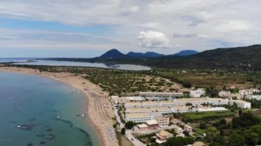 Aerial footage of the beautiful small town known as St George South, Greek city of Corfu Greece, showing people relaxing on the beach with hotels and mountains in the background on the Issos Beach