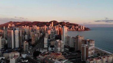 Aerial footage of Benidorm at sunset showing the beaches, hotels, and buildings with the sun reflexing off the apartment blocks.