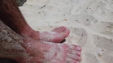 Güneşli bir günde, tropikal kumsalda kumla oynayan insanların ayakları okyanus kıyısında tatil yapan beyaz adamların ayak ve ayak parmaklarını gösteriyor..