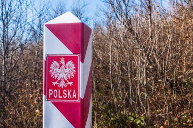Polish, white and red border post. On the pole, there is a white eagle with a golden crown and the inscription Polska - Poland. In the background, the blue sky and trees, bushes and grass.