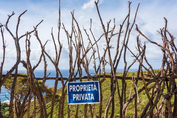 A wooden fence built of dry branches against the background of a green hill and blue sea. On the fence a blue sign with the Italian text \