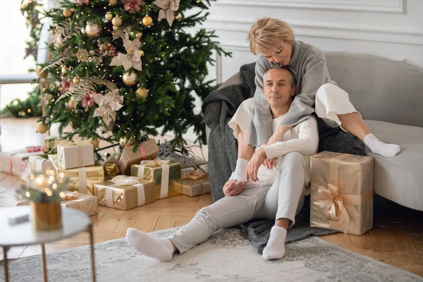 Yetişkin bir adam ve bir kadın Noel ağacının yanındaki kanepede oturur ve sarılırlar.