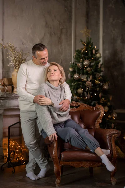 Güzel, yetişkin bir kadın ve bir adam bir koltuğa oturur ve bir Noel ağacı ve şöminenin yanında sarılırlar.