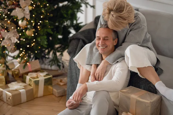 Yetişkin bir adam ve bir kadın Noel ağacının yanındaki kanepede oturur ve sarılırlar.