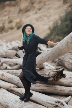 Mavi saçlı ve şapkalı güzel bir kadın rüzgarlı sahilde yürüyor.