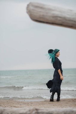 Mavi saçlı ve şapkalı güzel bir kadın rüzgarlı sahilde yürüyor.