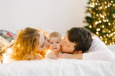 Mutlu anne ve baba Noel ışıklarının arka planında çocuklarının yanında yatar ve onu yanaklarından öperler.
