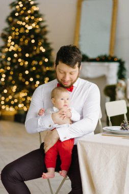 Beyaz gömlekli, kırmızı papyonlu şık bir baba oturma odasındaki Noel ışıklarına karşı oğlunu kollarında tutuyor.