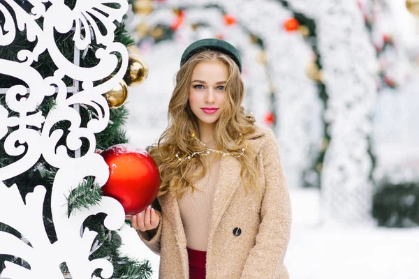 一个年轻时髦的女人 有一个红色的圣诞树大舞会 背景上是一个节日的圣诞市场 这个模特穿着时髦的冬衣 — 图库照片