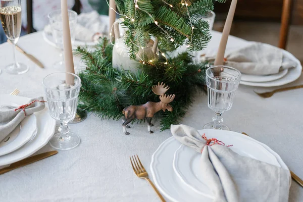 Christmas decor of the festive table: plates, napkins, cutlery, a statuette of an elk and fir branches in a vase with lights