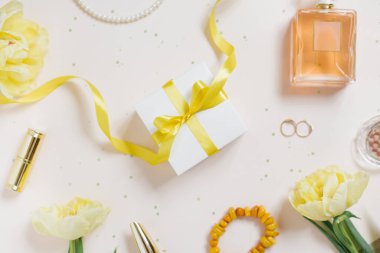 Doğum günü kartı: üzerinde sarı fiyonk ve kurdele olan hediye kutusu, sarı lale çiçekleri, kozmetik ürünleri ve açık bej bir masa üzerindeki kadın aksesuarları. Düz yatıyordu