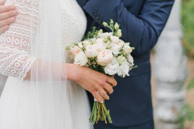 The bride holds in her hands a beautiful bouquet of delicate flowers and green leaves. The newlyweds are embracing, the hands of the bride and groom are close-up.
