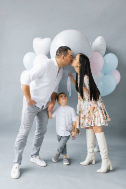 A young stylish family waiting for their second child is kissing against the background of a pop ball to determine the gender of the future baby. Parents with their eldest son on a gray background. Gender reveal party