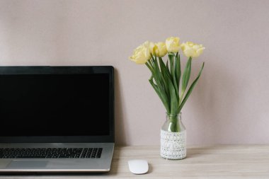 Spring home office still life composition. Blank laptop screen. Yellow tulips in a vase on the table. Office workplace. Scandinavian interior.