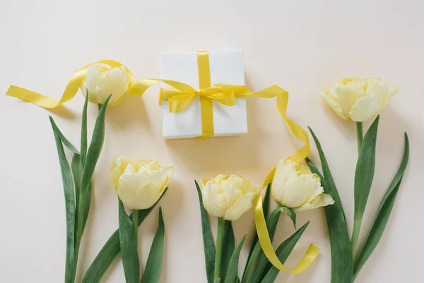 ギフトボックスは 薄いテーブルの上に黄色のリボンと黄色のチューリップの花で白い紙に包まれています — ストック写真
