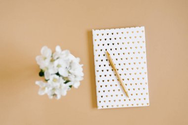 Kapağında altın kalpler olan beyaz bir defter, bir vazoda duran altın kalem ve bej bir masa üzerindeki beyaz elma çiçekleri. Düzen ve tasarım konsepti. Düz yatıyordu.