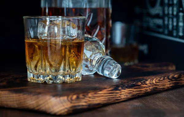 A glass of whiskey and ice on a wooden board against a dark background. — Fotografia de Stock