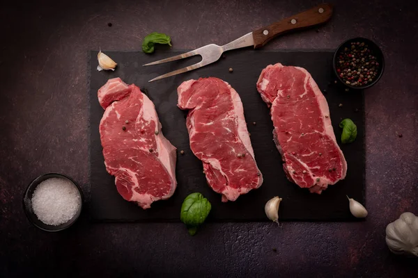 Top down view of raw striploin steaks on a black slate board, with seasoning. — Fotografia de Stock