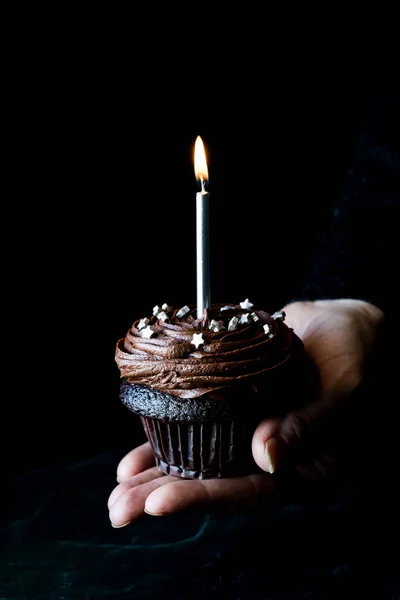 Mão segurando um cupcake de chocolate com uma vela acesa, contra um fundo preto. — Fotografia de Stock