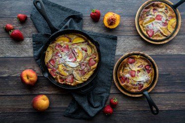 Strawberry and peach Dutch baby pancakes in frypans covered with powdered sugar, against a dark background.