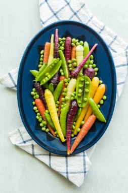 Top view of an oval dish of rainbow carrots, peas and pea pods, against a light background. 