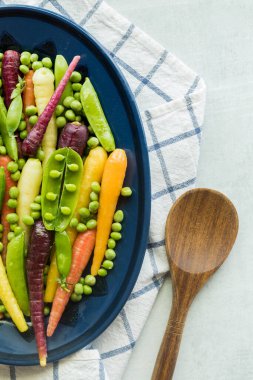 Top view of half of a dish of rainbow carrots with peas and pea pods, against a light background.
