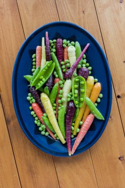 A colourful platter of fresh assorted peas and rainbow carrots, on a wooden table. 