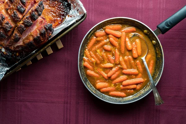 从上往下看油炸锅里的油酥胡萝卜 旁边的锅里放着一个有釉面的熏制野餐火腿 — 图库照片