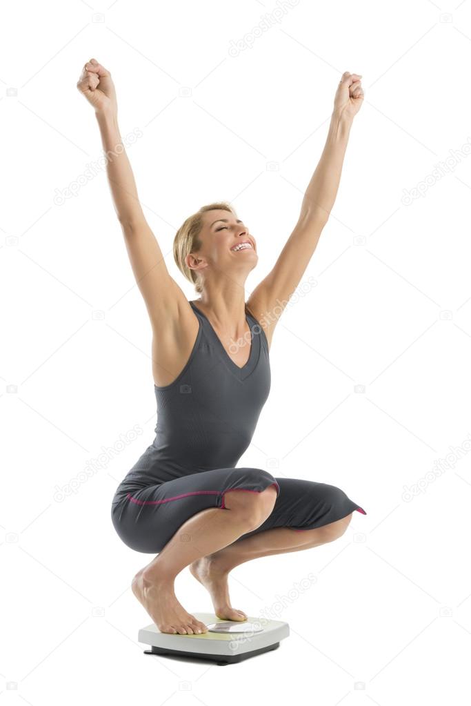 Happy Woman With Arms Raised Crouching On Weight Scale