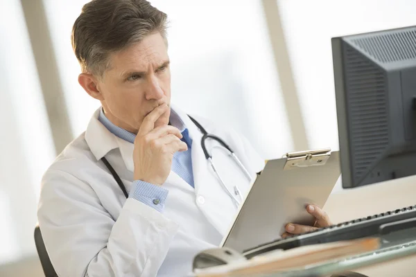 Doctor serio mirando a la computadora mientras sostiene el portapapeles en De — Foto de Stock