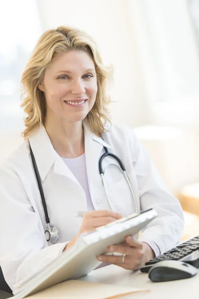 Female Doctor With Clipboard Sitting At Desk In Clinic — Stock Photo, Image