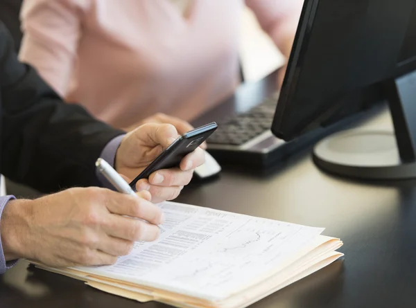 Empresário usando telefone inteligente enquanto escreve no documento na mesa — Fotografia de Stock