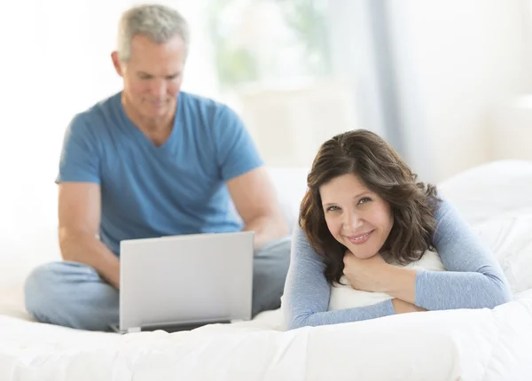 Žena v posteli, zatímco muž doma pomocí přenosného počítače — Stock fotografie
