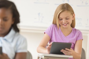 Schoolgirl Smiling While Using Digital Tablet At Desk clipart