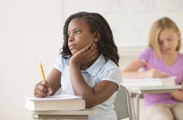 Chica reflexiva con la mano en la barbilla sentada en el escritorio del aula —  Fotos de Stock