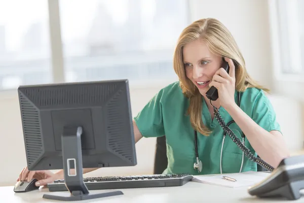 Médico usando la computadora mientras conversa en el teléfono fijo en el escritorio —  Fotos de Stock