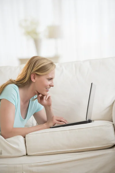 Mujer feliz usando el ordenador portátil mientras está acostado en el sofá —  Fotos de Stock