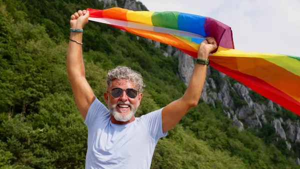 Gri saçlı, yaşlı, biseksüel, sakallı, güneş gözlüklü, doğada LGBTQIA bayrağı taşıyan yaşlı bir adamın portresi. Onur Ayı 'nı, Gökkuşağı Bayrak Günü' nü, gay geçit törenini kutluyor