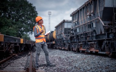 Mühendis demiryolu inşaat sürecinde petrol kargo treni ve tren istasyonundaki demiryolu çalışmalarını tabletle kontrol ediyor. Mühendis iş yerinde güvenlik üniforması ve güvenlik kaskı takıyor..
