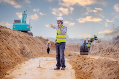 Güvenlik üniforması, miğfer ve telsiz iletişimi giyen mühendisler, teodolit ekipmanlarıyla yolun inşaat makineleri arka planına sahip inşaat sahasındaki ölçüm sahasında konumlandırılmaları için teodolit kullanıyorlar..