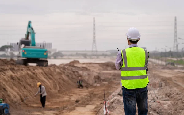 Construction engineer wear safety uniform under inspection and survey workplace by tablet with excavation truck digging and worker construction road background