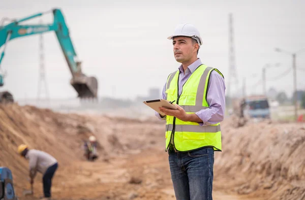 Construction engineer wear safety uniform under inspection and survey workplace by tablet with excavation truck digging and worker construction road background