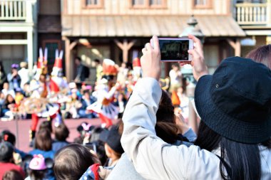 Siyah şapkalı bir kız kalabalığın yanında duruyor ve telefonuyla geçit töreninin fotoğrafını çekiyor. 
