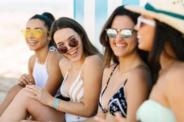 Four young women in bikinis laughing with each other leaning against the beach wall.