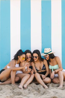 Four young women in bikinis using smart phones leaning against the beach wall